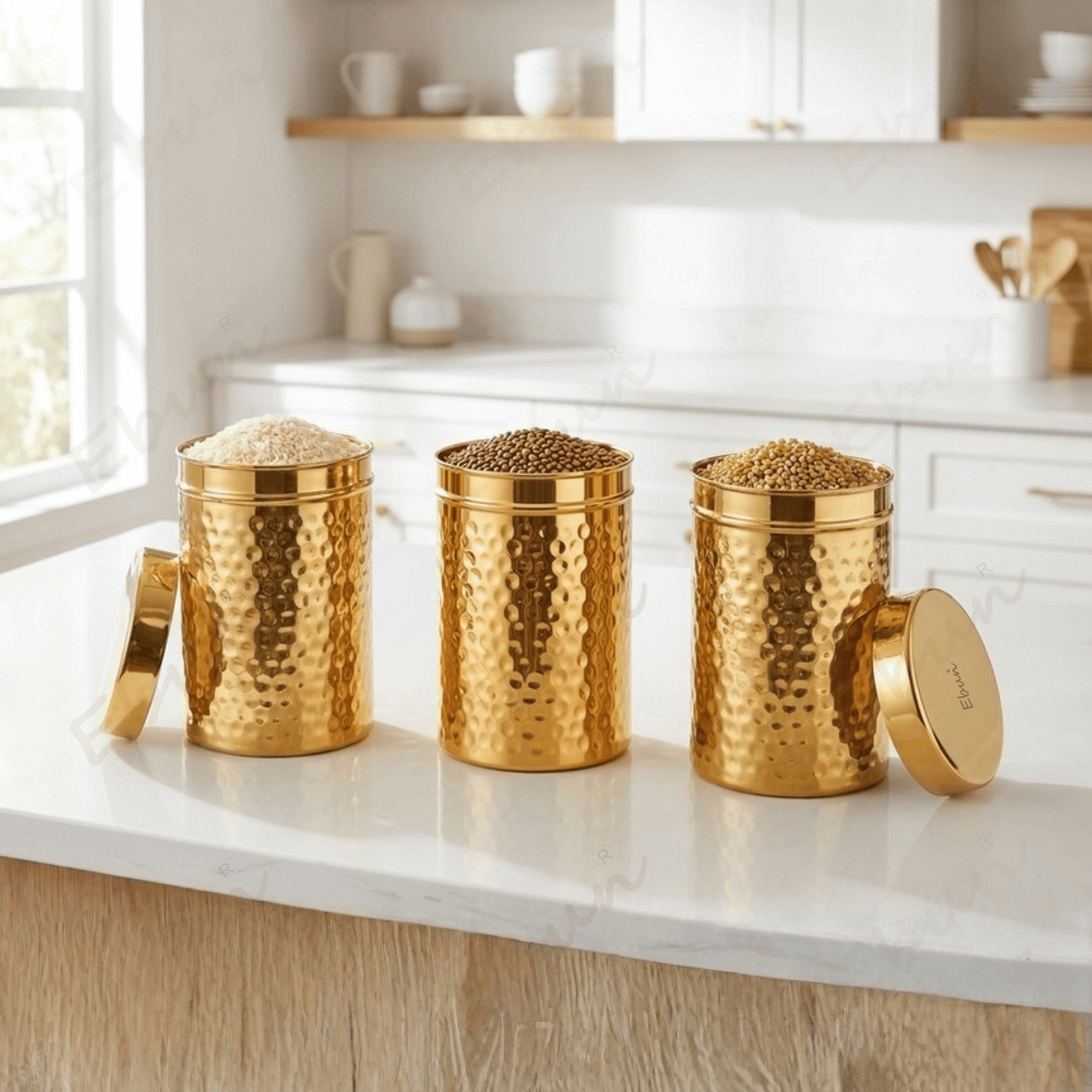 Three hammered gold metal canisters with matching lids sit on a white kitchen countertop, filled with grains or seeds, in a bright modern kitchen setting.