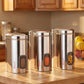 Three Ebun stainless steel kitchen canisters on a wooden countertop with a blurred background of a kitchen.
